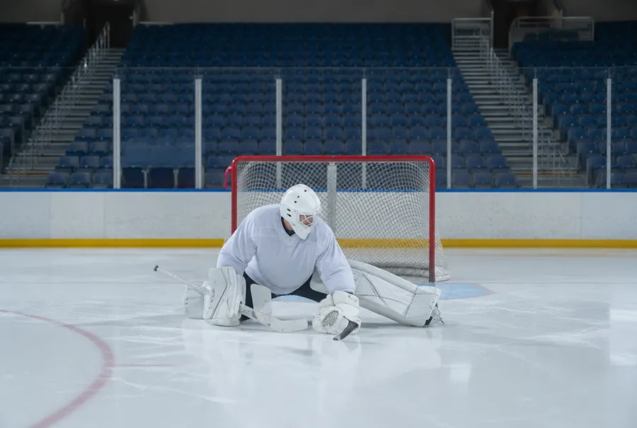 Hockey goalie in plain gear stretching alone on the ice of an empty arena before morning practice